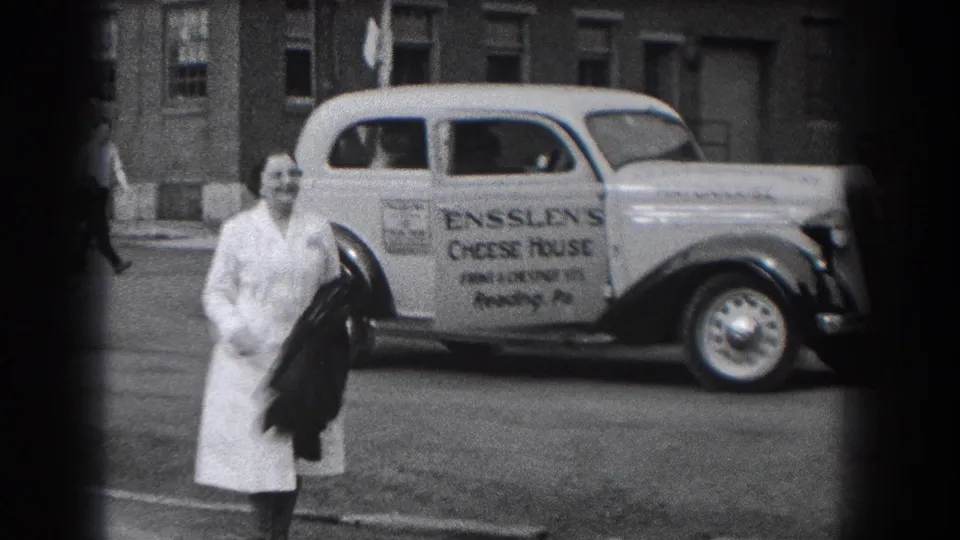 A woman in a light dress walks toward the camera while the Ensslen's Cheese House delivery car remains parked behind her.