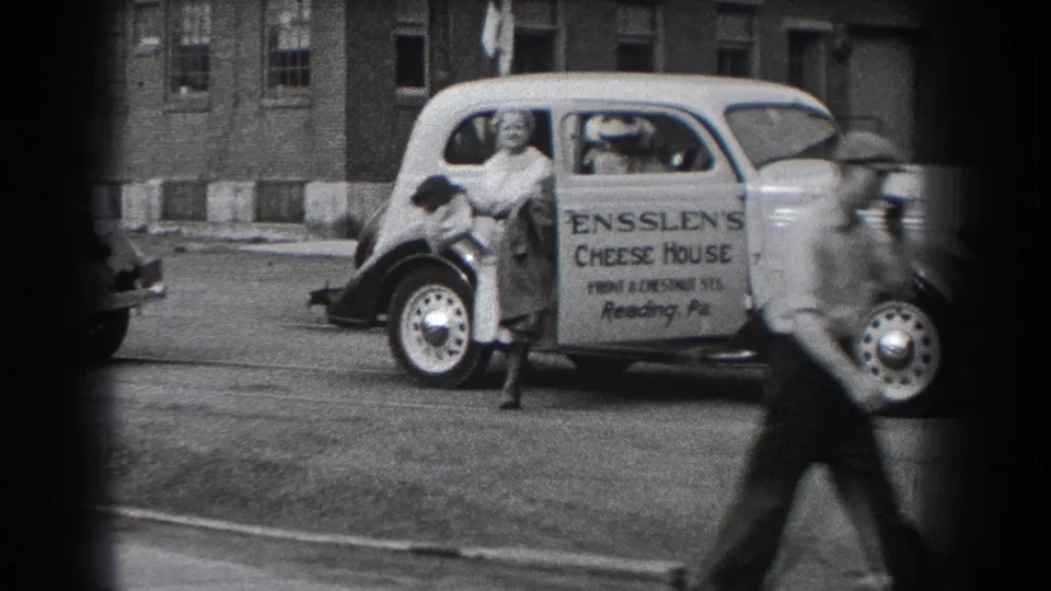 A woman near the Ensslen's Cheese House car is partly obscured as a man in a hat crosses the foreground on the Reading street.