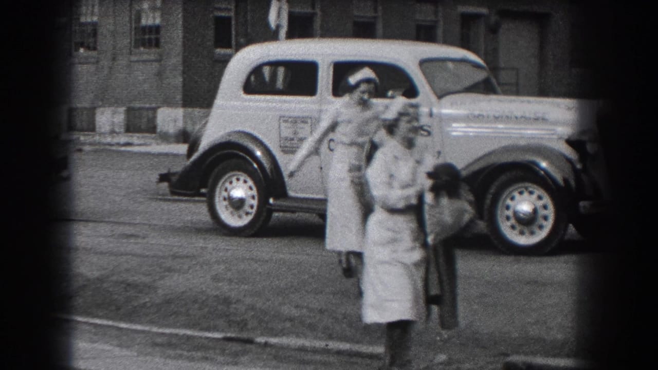 Two nurses carrying coats walk in front of a light-colored car with side lettering.