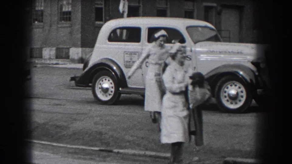 Two nurses carrying coats walk toward the camera beside a lettered delivery car in Reading, Pennsylvania, in 1938.