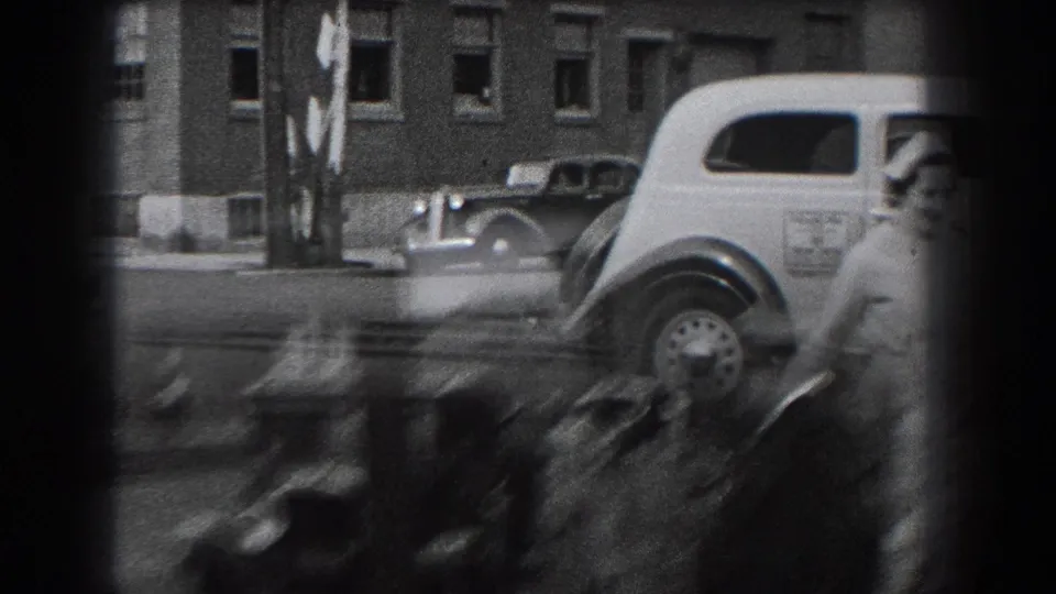 A nurse exits the frame near the light-colored delivery car while another vintage car passes in the 1938 Reading street scene.