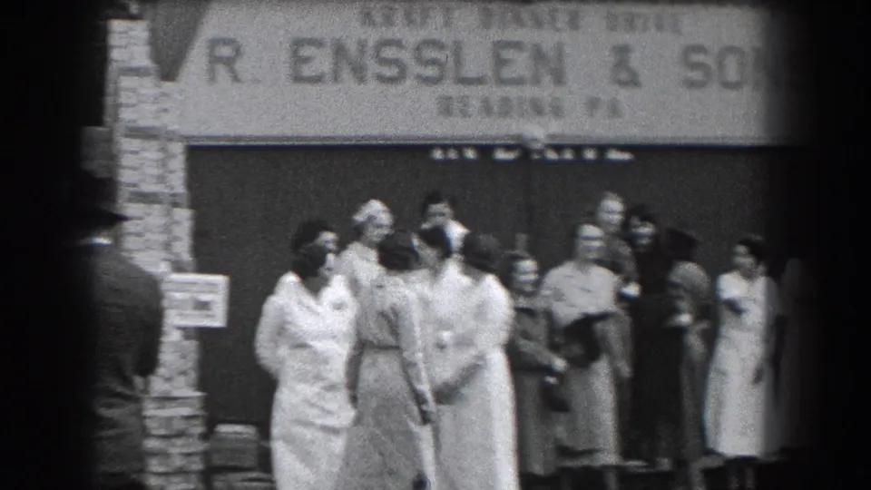 A small group stands near the R. Ensslen & Sons sign in Reading, Pennsylvania, with uniformed women in the foreground and suited men behind them.