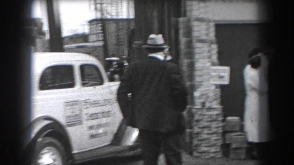 In Reading, Pennsylvania, a suited man in a fedora passes an Ensslen's Cheese House delivery car outside a commercial doorway.