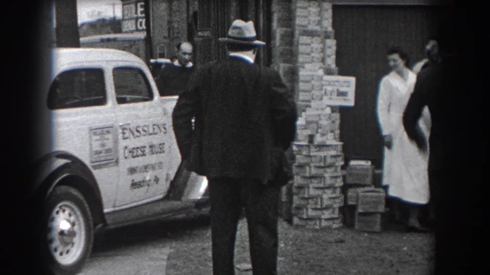 A woman in a white coat stands at the edge of the Reading storefront scene, with the Ensslen's Cheese House car and stacked boxes nearby.
