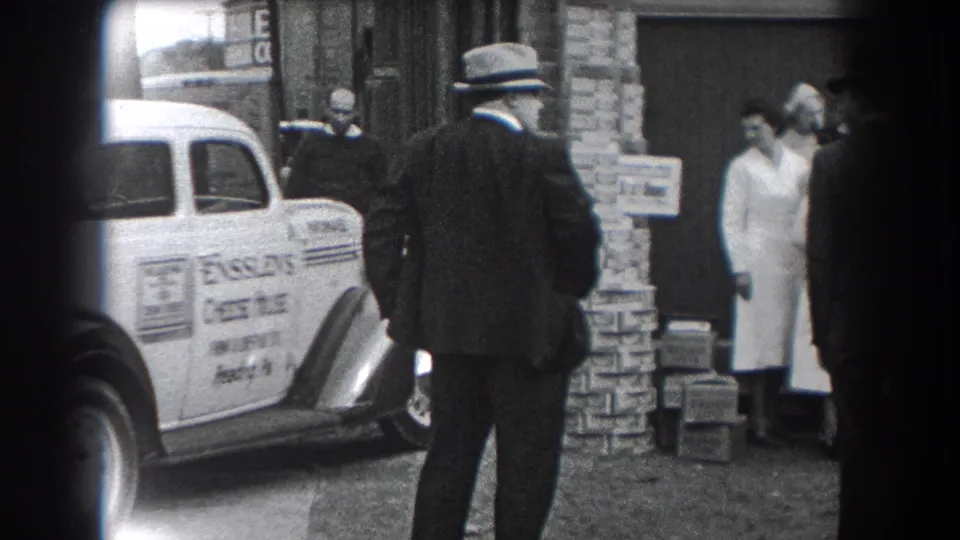In 1938 Reading, Pennsylvania, a suited man faces a white Ensslen's Cheese House delivery car as people stand near a brick storefront.