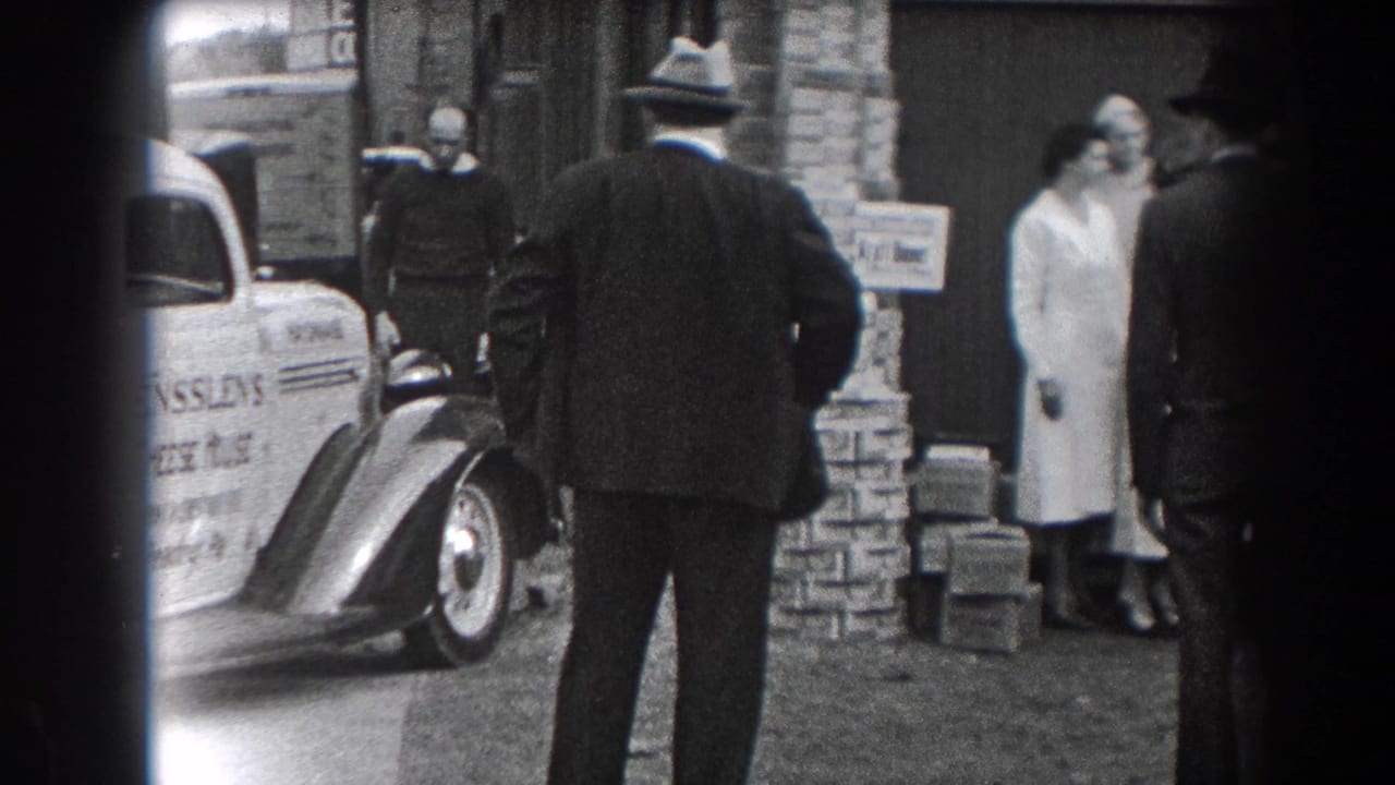 A fedora-wearing man stands with his back to the camera near a parked delivery car.