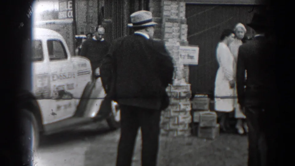 The home-movie frame shows a suited man, women in light coats, boxes, and a marked cheese delivery car outside a Reading brick building.