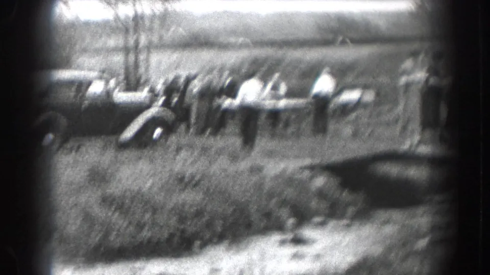 A blurred 1938 home-movie frame shows parked automobiles in an outdoor lot or street area, with strong grain and dark frame edge visible.