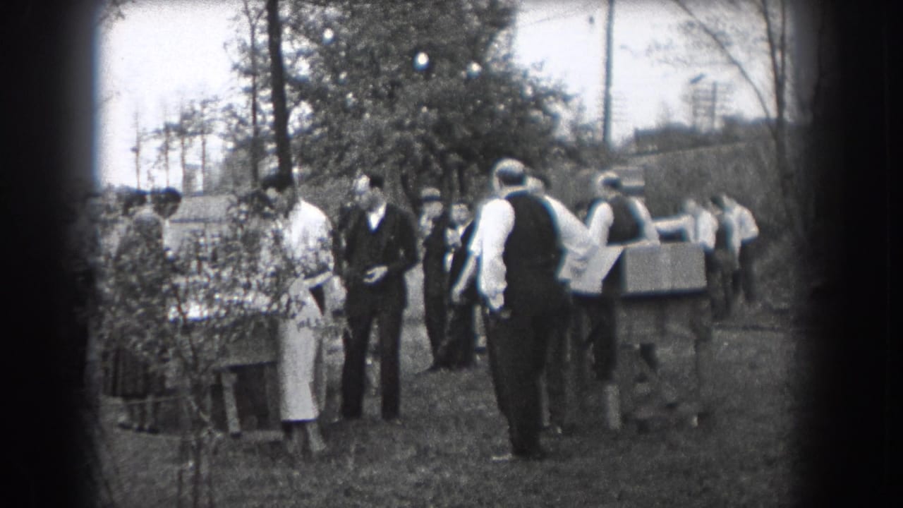 Adults in period clothing stand and talk near shrubs, a table, and trees in 1938 Reading, Pennsylvania footage.