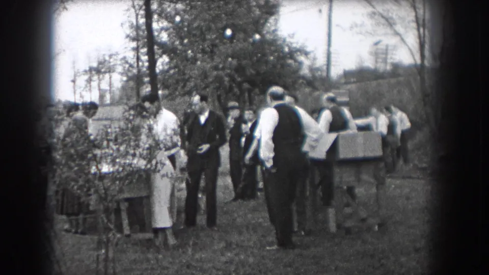 Adults in period clothing stand and talk near shrubs, a table, and trees in 1938 Reading, Pennsylvania footage.