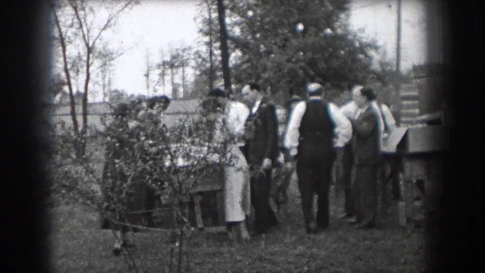 A home-movie frame shows adults gathered beside bushes and a table in a grassy Reading, Pennsylvania outdoor setting.