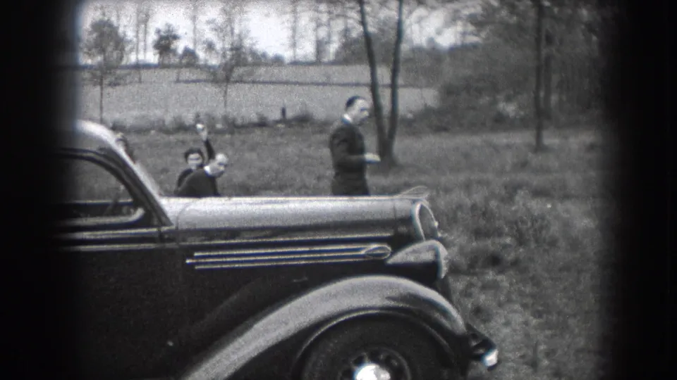 Black-and-white 8mm frame of a child greeting the camera near a parked automobile, with an adult man standing in a grassy Reading, Pennsylvania setting.