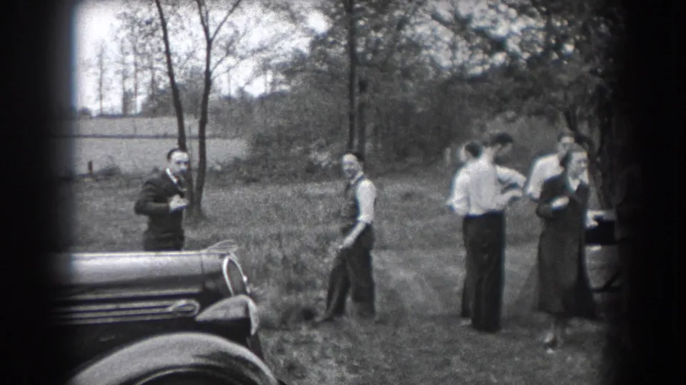 Adults in 1930s attire appear to converse and move around a lawn near a parked car in Reading, Pennsylvania.