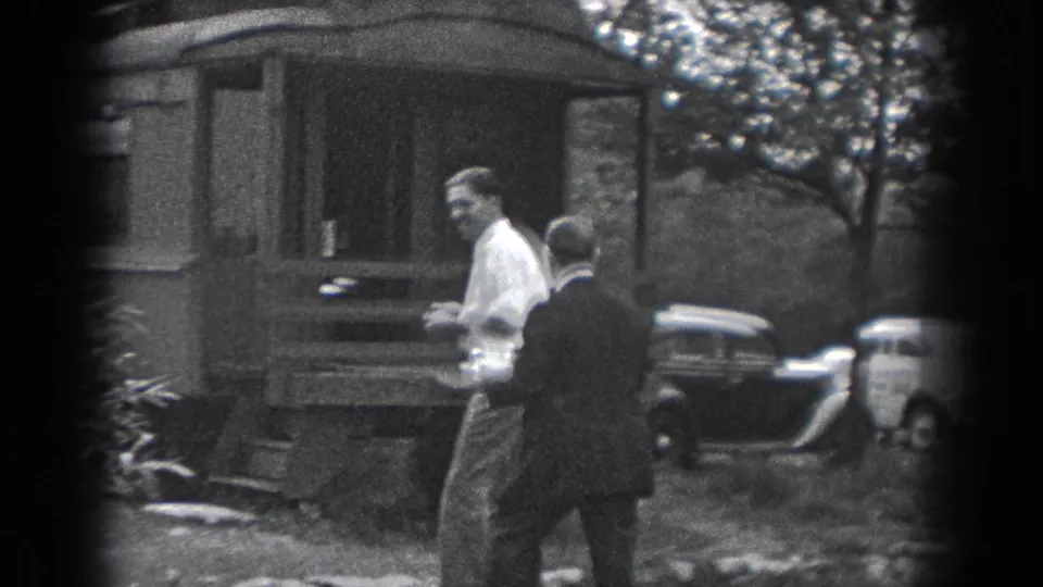 Two men in period clothing stand near a small outbuilding while cars are parked in the background in Reading, Pennsylvania.