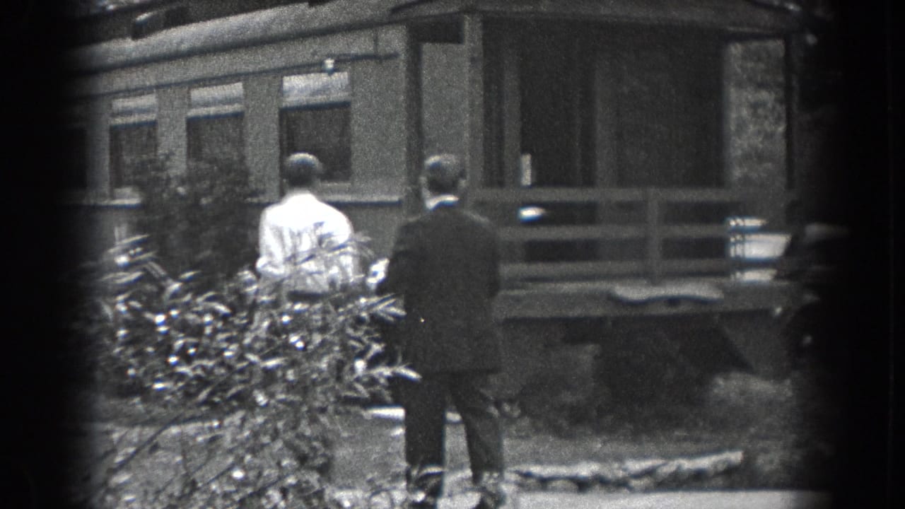 Two adult men in 1938 Reading, Pennsylvania walk toward a modest porch, with shrubs and a parked car in the yard view.