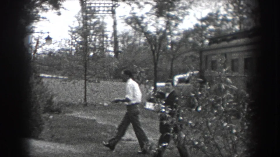 A man in a light shirt crosses the yard in Reading, Pennsylvania, with a modest house, trees, and parked period cars visible.