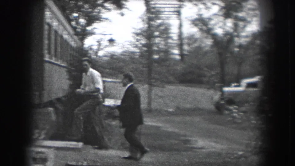 Two adult men walk beside a house in 1938 Reading, Pennsylvania, with trees, grass, sidewalk, and a parked period car in view.