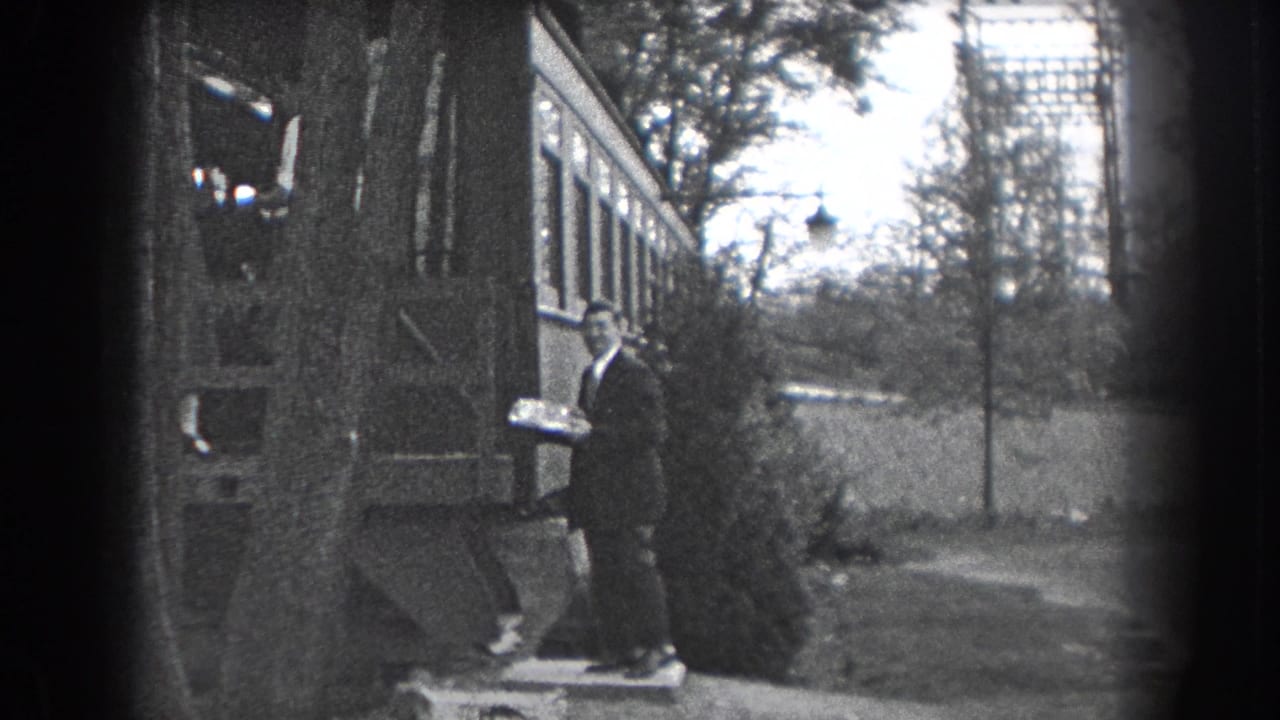 Man in a dark suit holds a cake or tray beside the house and garden.