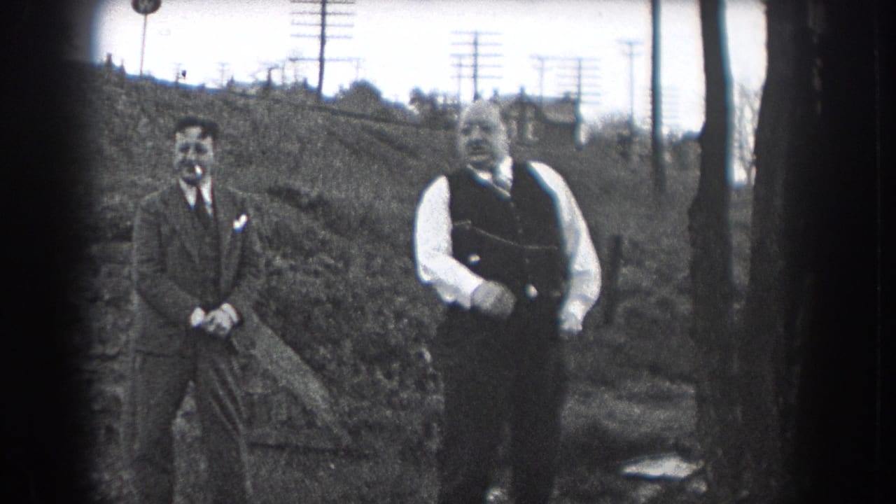 Two men in suits stand outdoors while one gestures with his arm.