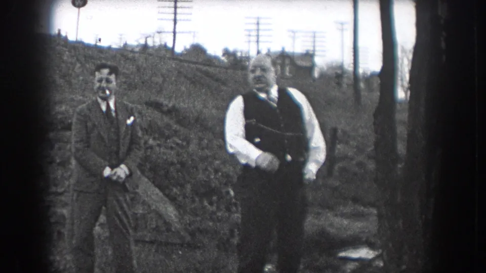 Two men in formal daywear stand outside in Reading, Pennsylvania, in 1938, with one man in a dark vest gesturing as the other watches near a sloped background.