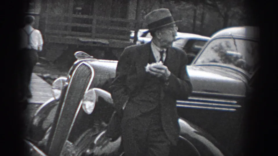 A man in a fedora and suit stands beside a vintage automobile in Reading, Pennsylvania, in 1938, holding a small object near his mouth as another figure appears in the background.