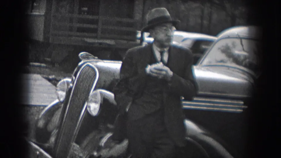 A suited man in a fedora stands beside the front of a vintage automobile in Reading, Pennsylvania, in 1938, with the car grille and rounded body visible behind him.