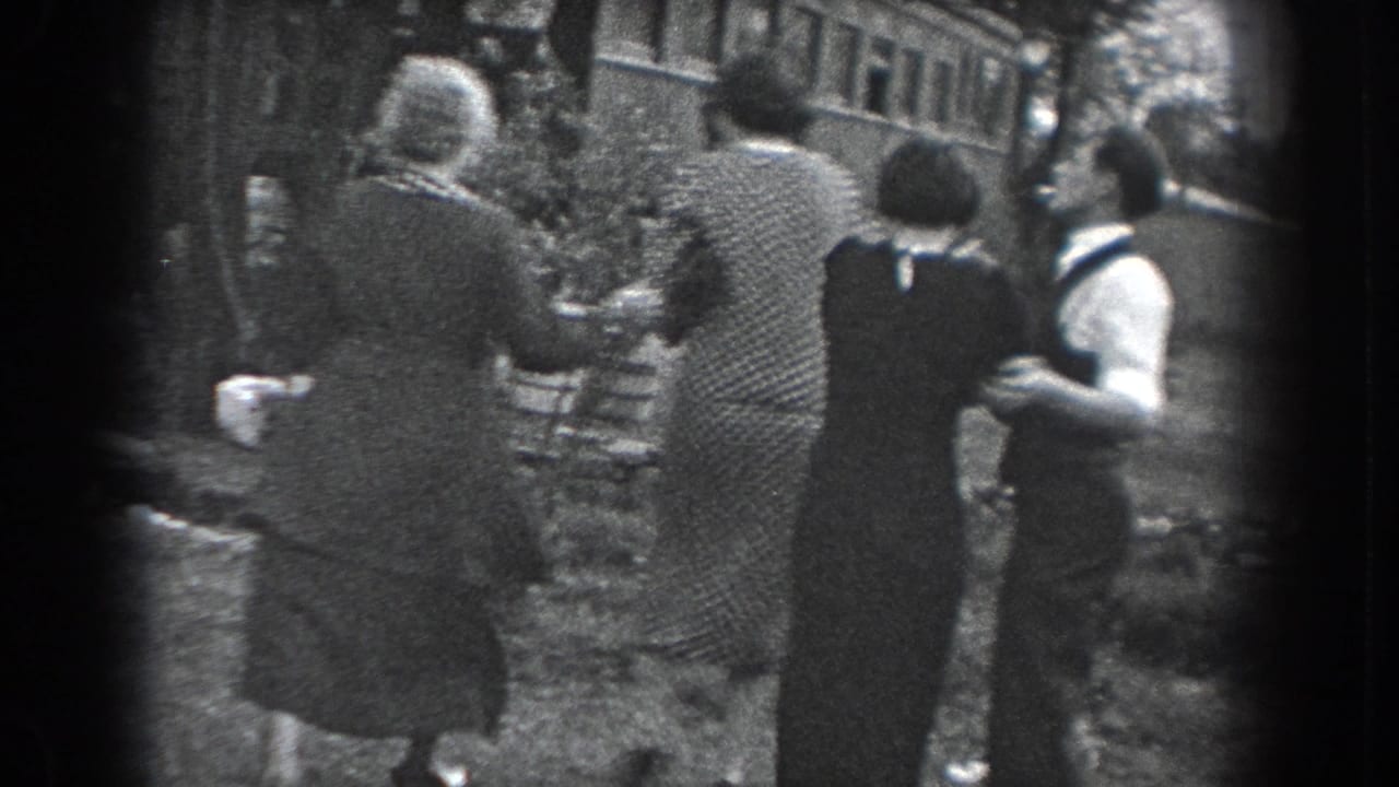 In 1938 Reading, Pennsylvania, several adults in period clothing walk away from the camera along a grassy path near a stone wall.