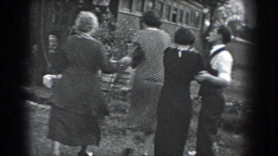 In 1938 Reading, Pennsylvania, several adults in period clothing walk away from the camera along a grassy path near a stone wall.