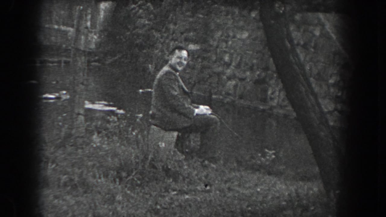 A man sits near water with trees and a stone wall behind him.