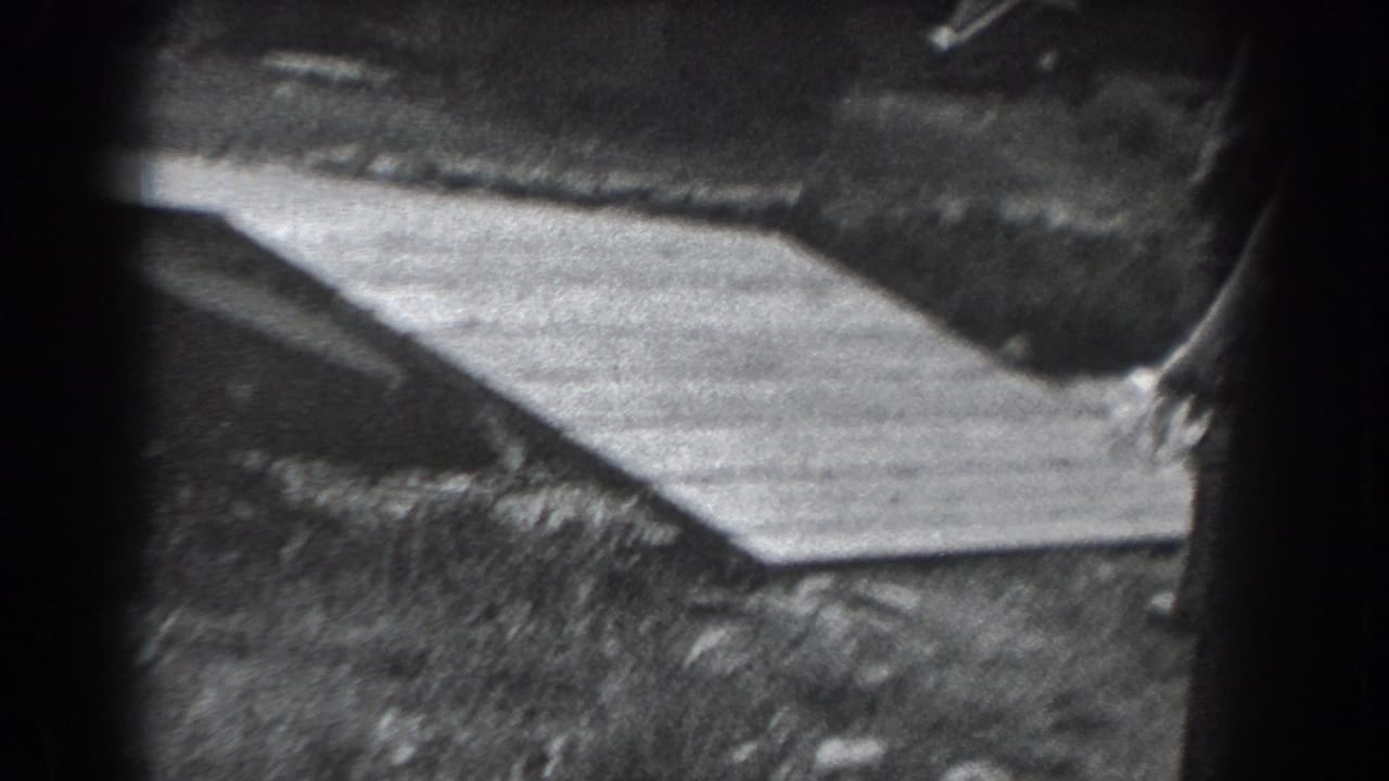 Dark 8mm frame with a wooden ramp, trees, and partial figures at the edge of the scene.