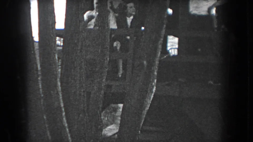 Grainy 1938 Reading, Pennsylvania home-movie frame showing people partially visible behind trees beside a rustic structure.