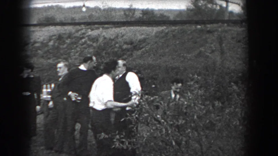 In 1938 Reading, Pennsylvania, adults in period clothing stand and talk near an outdoor table by a grassy hillside.