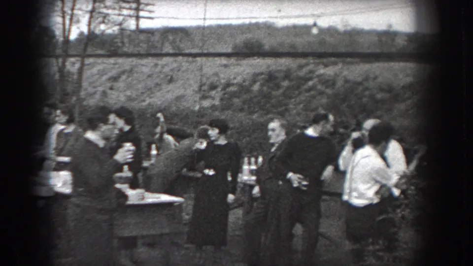 Adults at a 1938 Reading outdoor gathering stand around tables with drinks, framed by trees and a grassy bank.