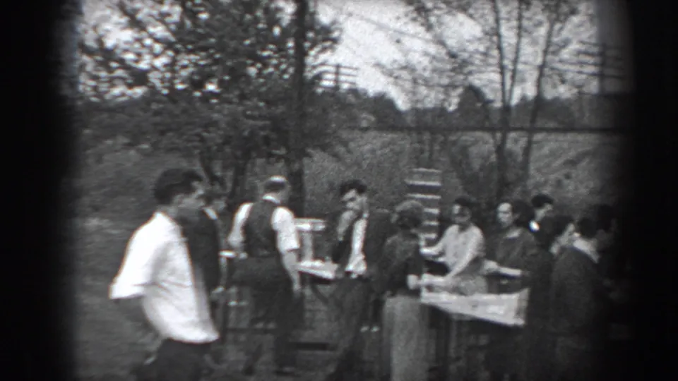 The home-movie frame shows adults gathered around food and drink tables in a wooded outdoor area in Reading, Pennsylvania.