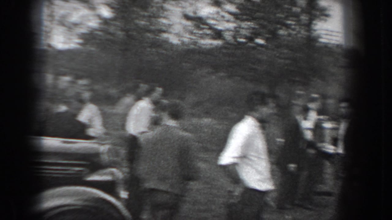 Men in suits gather on grass near a dark car in a wooded outdoor area.