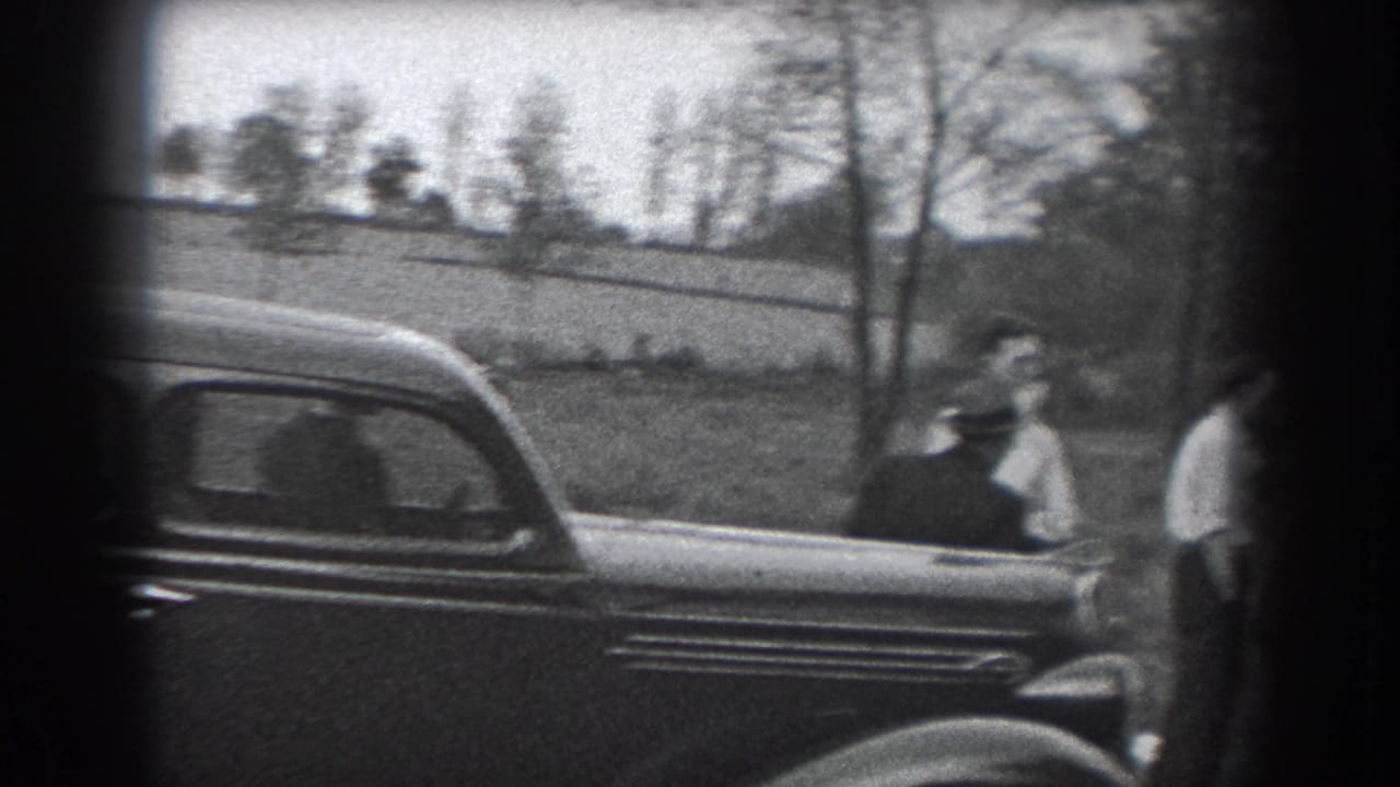 A dark classic car is parked beside men standing in a grassy area.