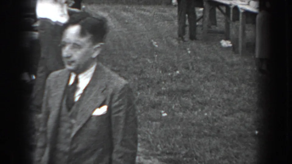 Foreground man in a suit stands on grass while picnic tables and other attendees appear behind him.