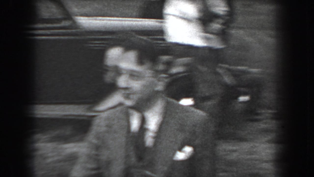 Suited man smiling near a parked automobile in soft black-and-white footage.
