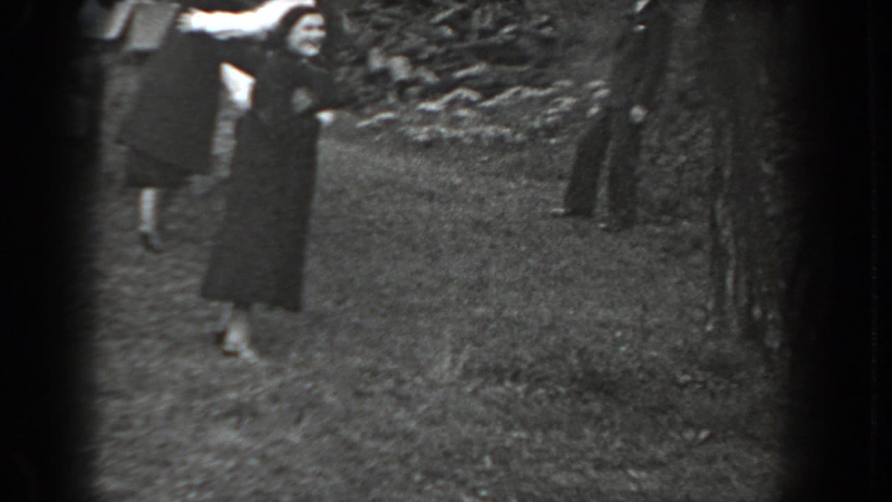 Woman in a long dark coat stands on grass with arms raised while adults watch nearby.