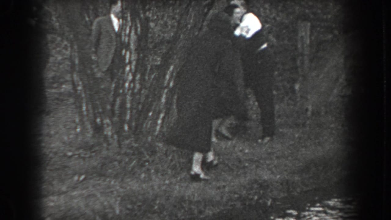 Woman in a dark dress pursues a suited man among trees while another man stands behind.