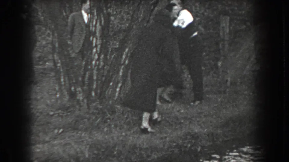 In the wooded Reading, Pennsylvania scene, a woman in a dark dress follows a suited man as a second suited man stands near the trees.