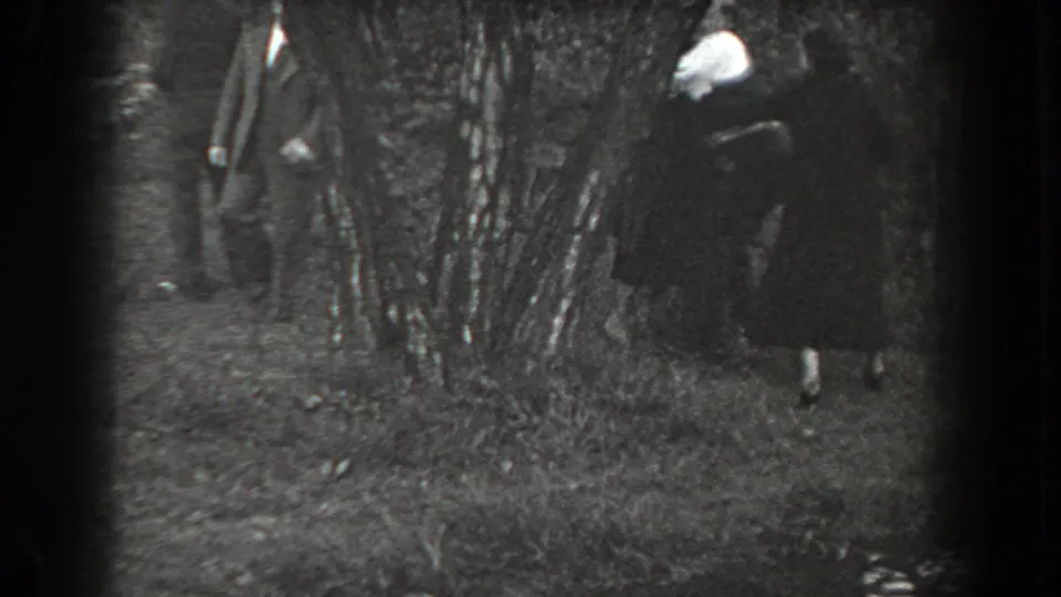 The home movie shows two suited men and a woman in a dark dress moving around trees in a grassy outdoor area.