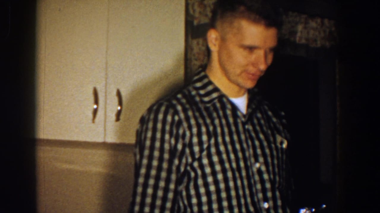 Young man in a checkered shirt standing in a dim kitchen.