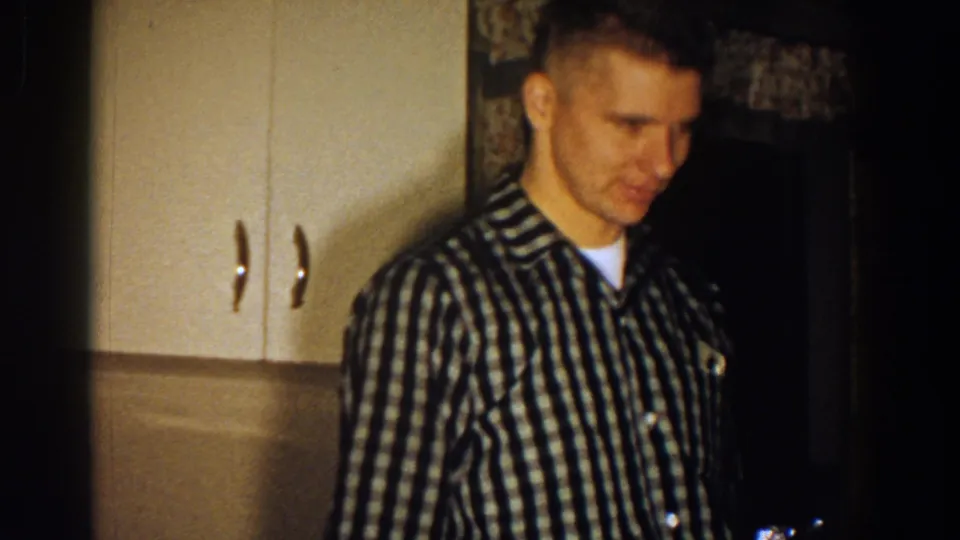 In 1958 Aliquippa home-movie footage, a young man in a checkered shirt stands near kitchen cabinets and floral curtains.