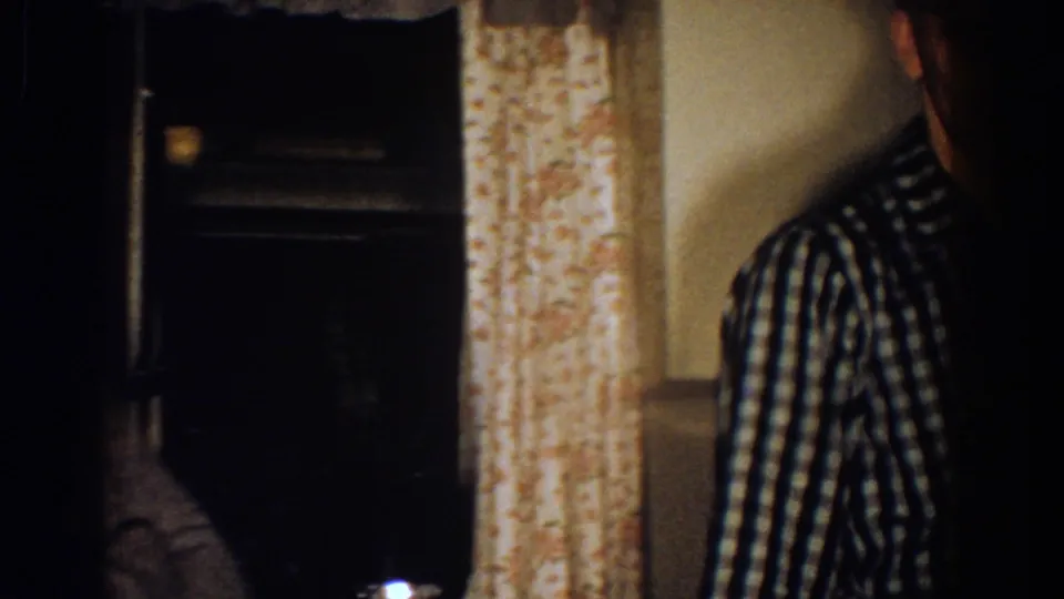 A partially obscured view shows the checkered-shirted man beside floral curtains in the dim 1958 kitchen interior.