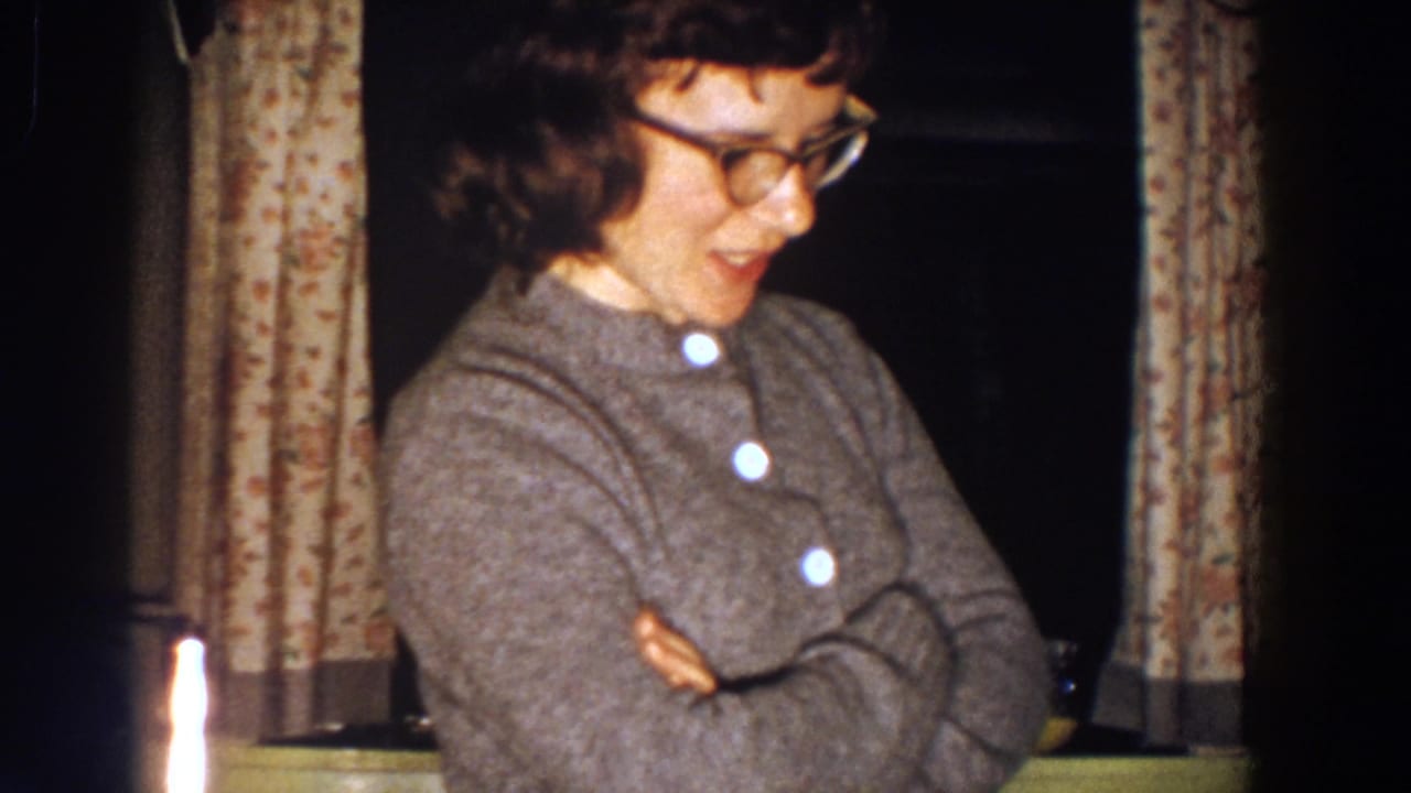 In 1958 Aliquippa home-movie footage, a woman in glasses and a grey cardigan stands with crossed arms near floral curtains.