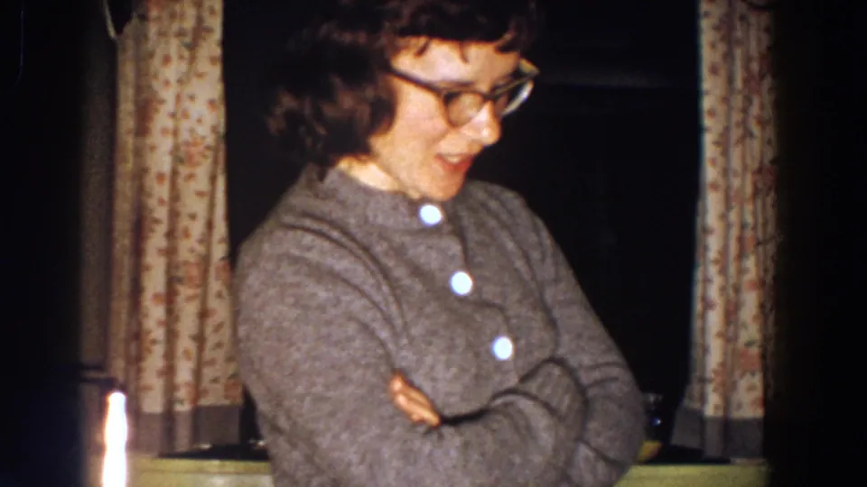 In 1958 Aliquippa home-movie footage, a woman in glasses and a grey cardigan stands with crossed arms near floral curtains.