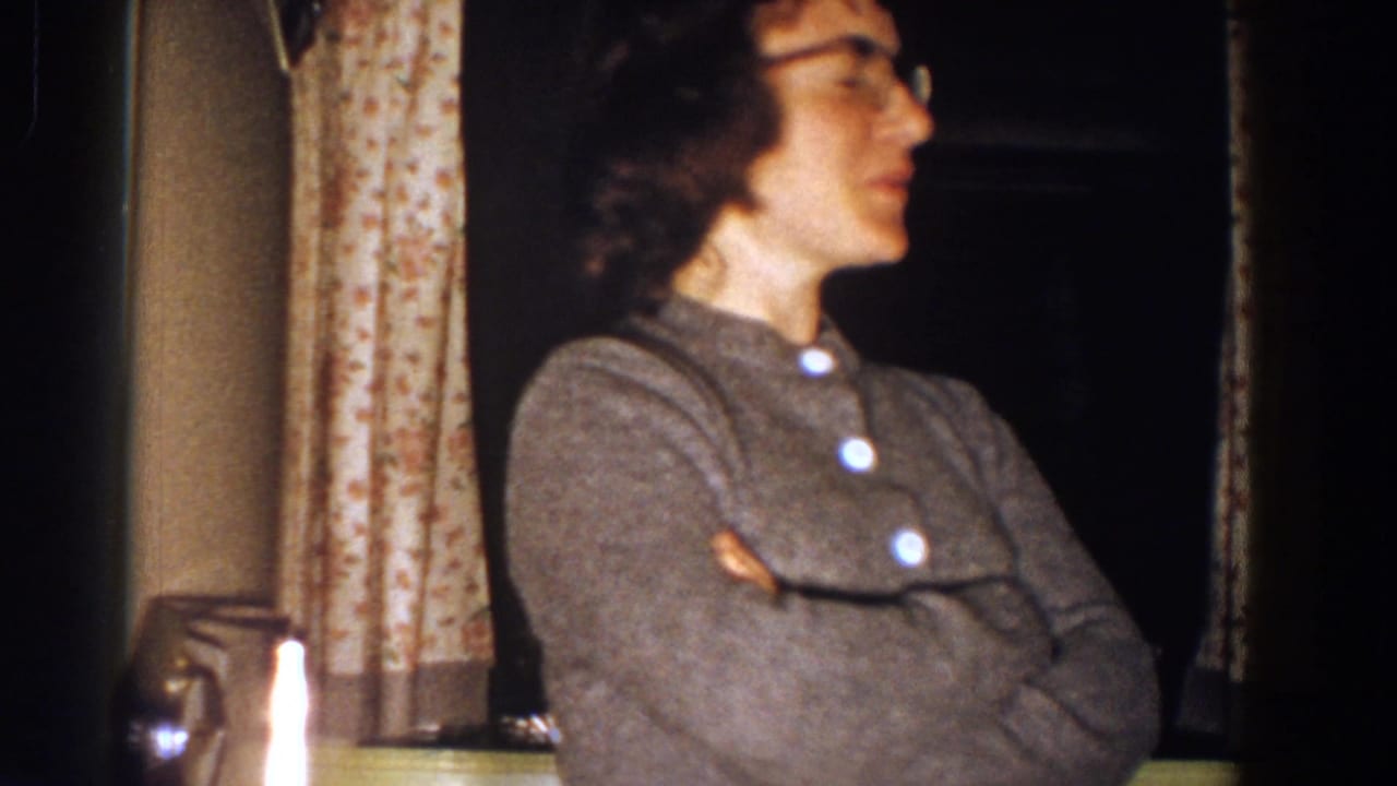 Woman in profile with crossed arms speaks near curtains and a dark window.