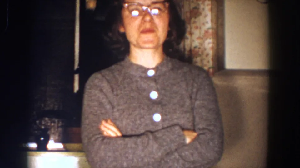 The woman faces the camera with arms crossed, glasses catching light, inside the Aliquippa home-movie setting.