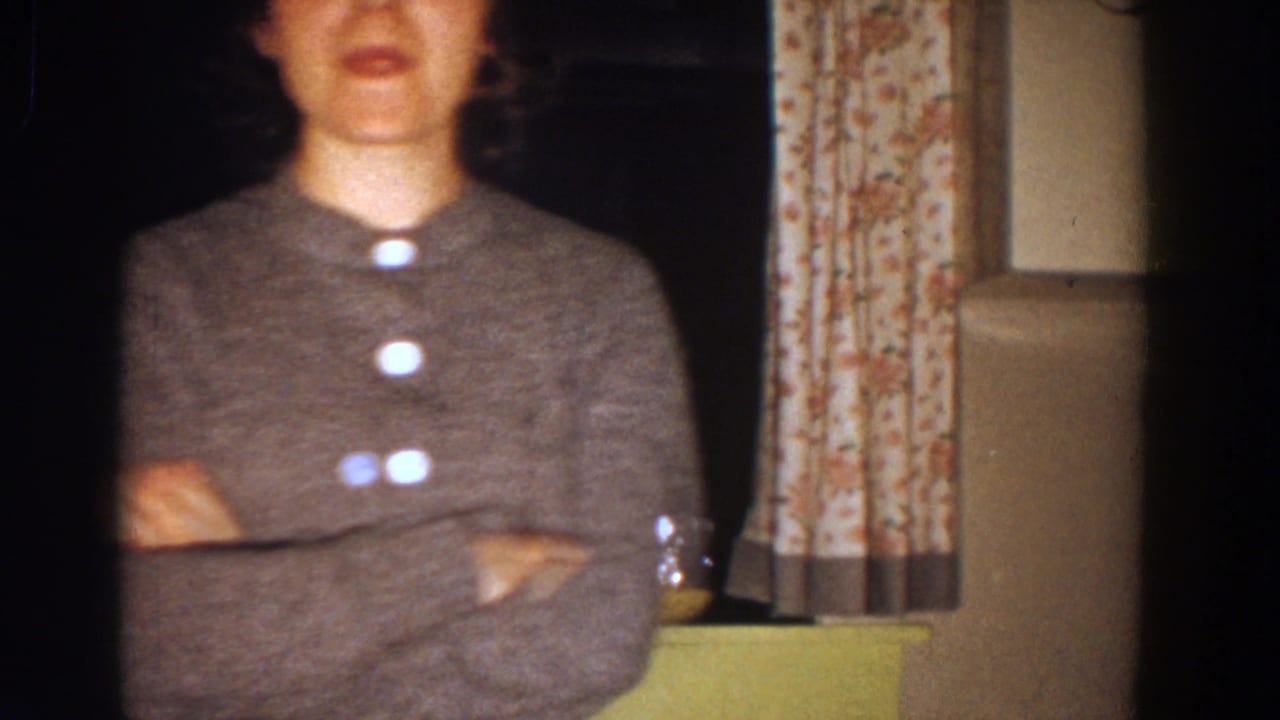 Woman in a gray sweater stands with arms crossed near floral curtains in a dim room.